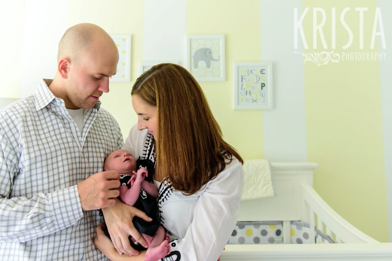 mother and father looking down at newborn baby girl while standing in yellow white and gray nursery during lifestyle family portrait session in their home
