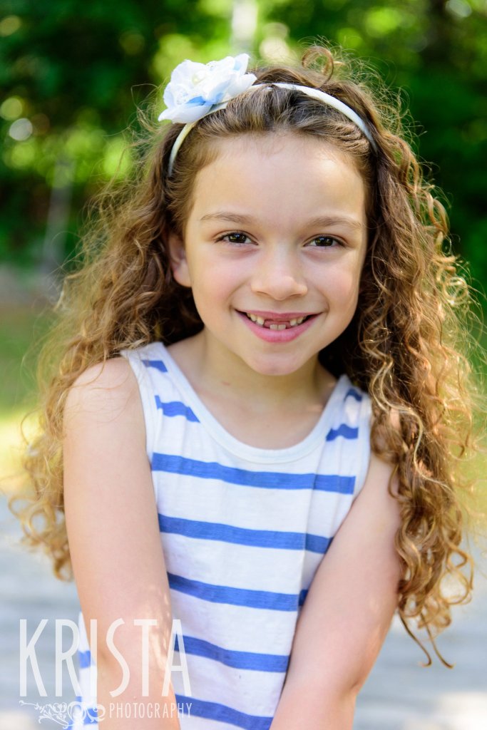 little curly haired girl in blue and white stripes sitting among green trees during springtime mini portrait sessions in New England