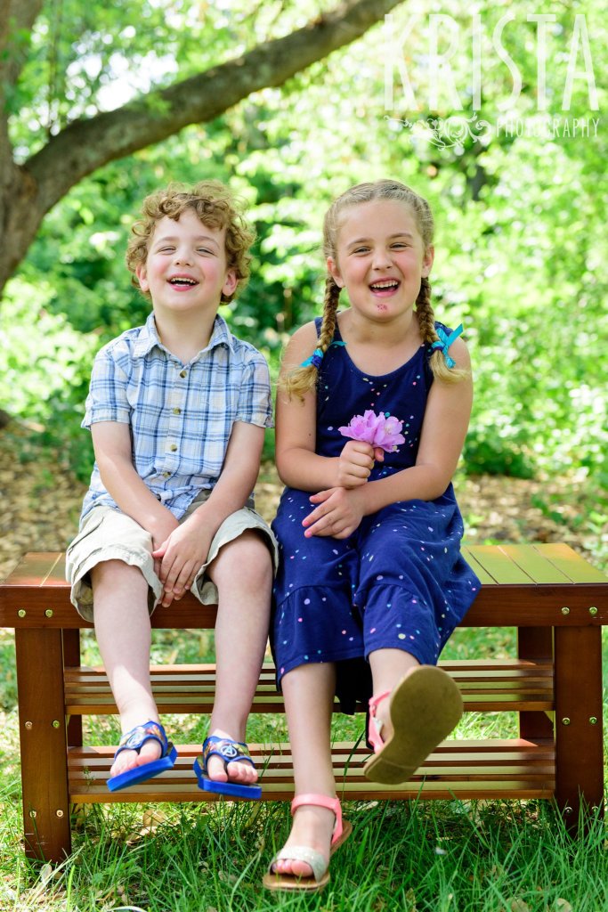 young brother and sister sitting on wooden bench laughing among green trees during springtime mini portrait session in New England