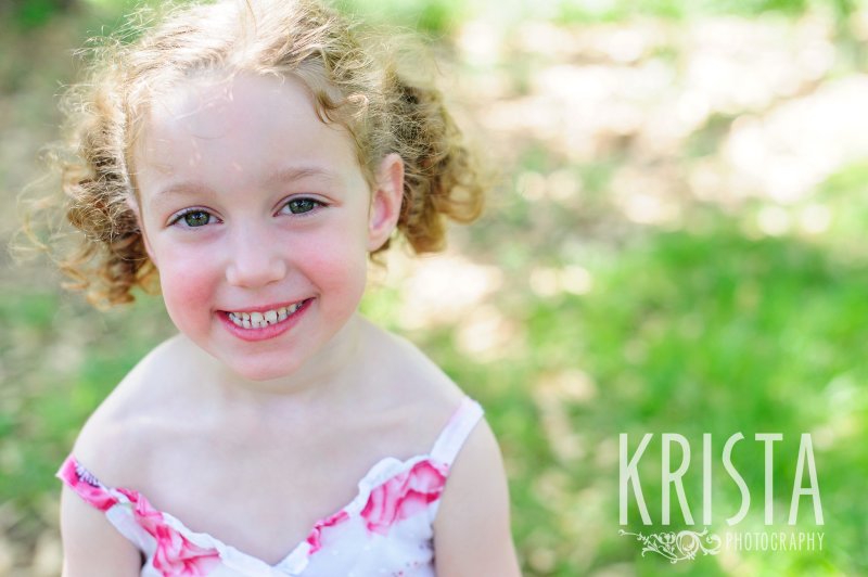 adorable little girl with curly hair in two pigtails in floral dress among green grass during spring mini portrait session