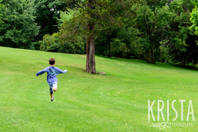 young boy running with arms out through green field during lifestyle family portrait session