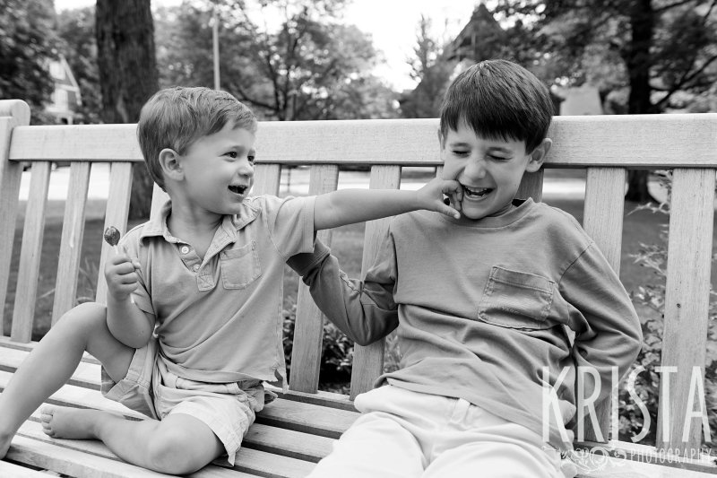 black and white brothers sitting side by side on outdoor bench younger boy punching at older boy who is laughing