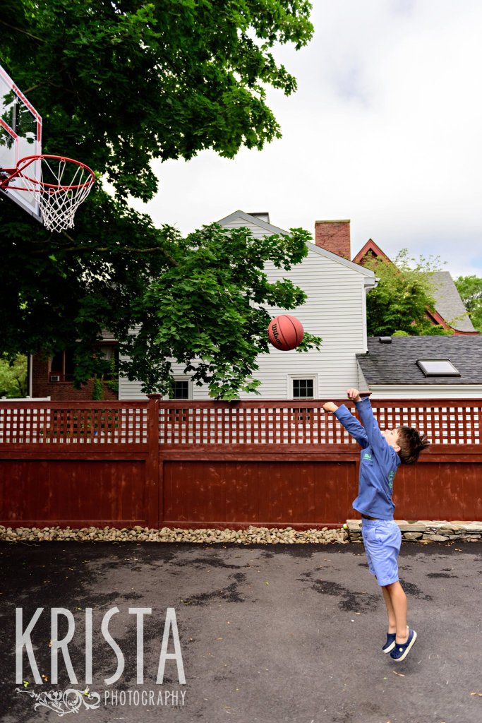 young boy in blue shooting basketball at hoop on driveway of home