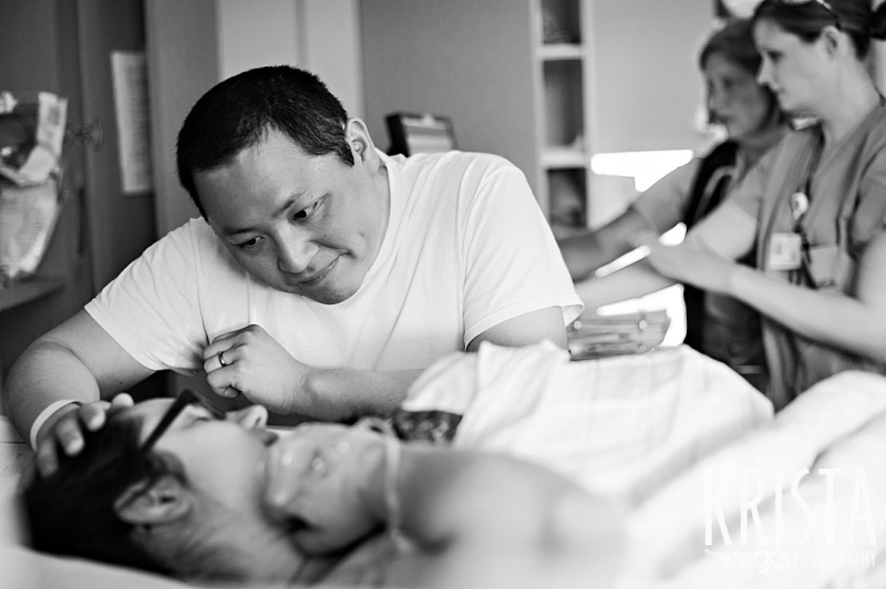 black and white image of father looking down at new mother holding newborn baby boy in hospital during lifestyle portrait session. Photo by Krista Guenin | Krista Photography
