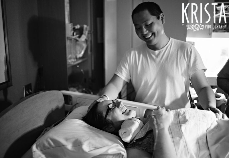 black and white image of father looking down at new mother holding newborn baby boy in hospital during lifestyle portrait session