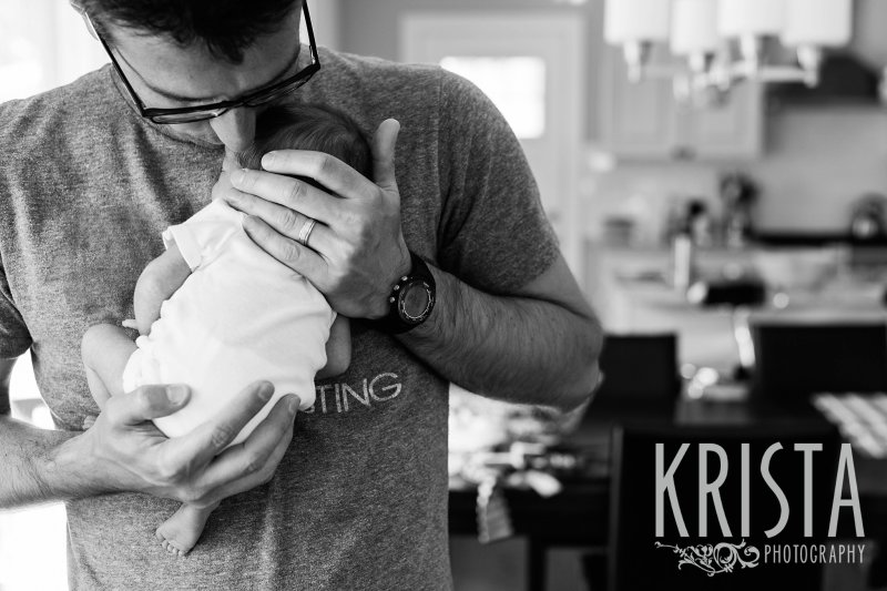 black and white image of father kissing newborn baby on head at home 
