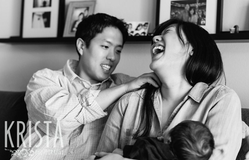black and white image of new parents laughing while holding newborn baby boy during portrait session in their home