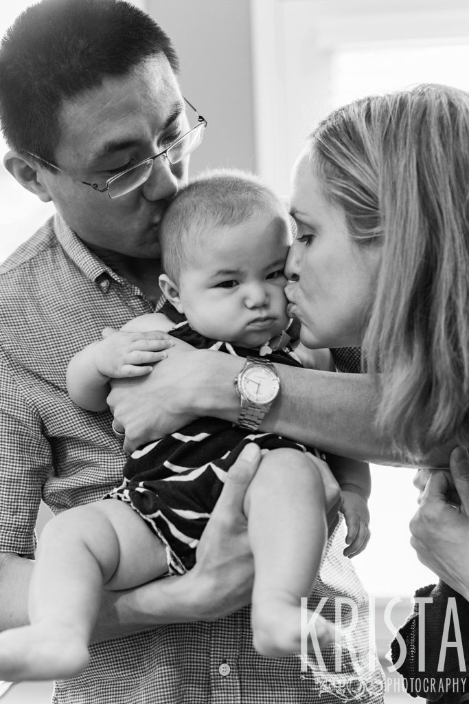 black and white image of new parents kissing baby girl who is not happy to be awake on the cheek during lifestyle portrait session at home