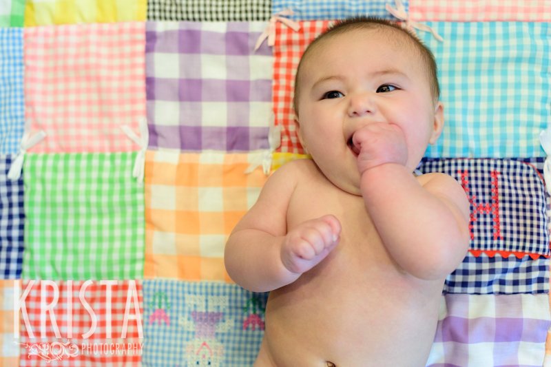 naked baby girl on quilt sucking hand and smiling during lifestyle portrait session at home
