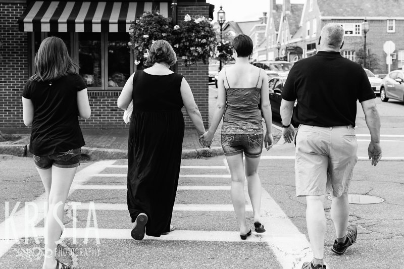 black and white image of family walking hand in hand across street during lifestyle portrait session near their home