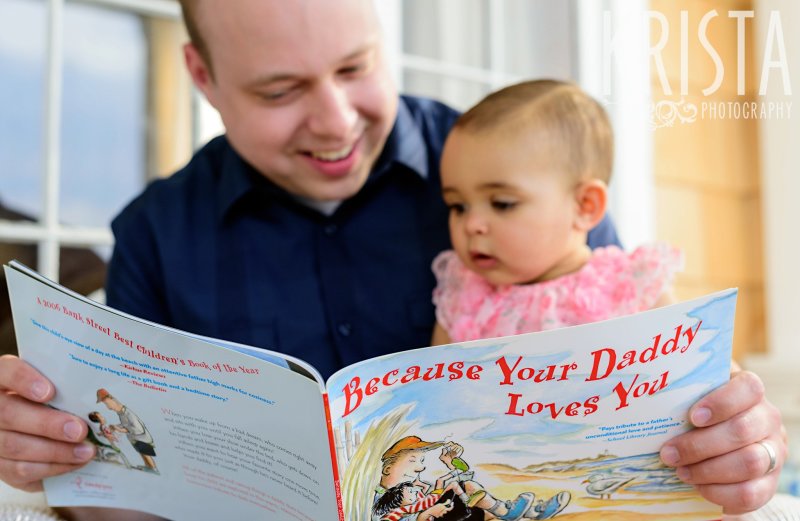 adorable baby girl on dad's lap reading "Because Your Dad Loves You" during lifestyle portrait session at home