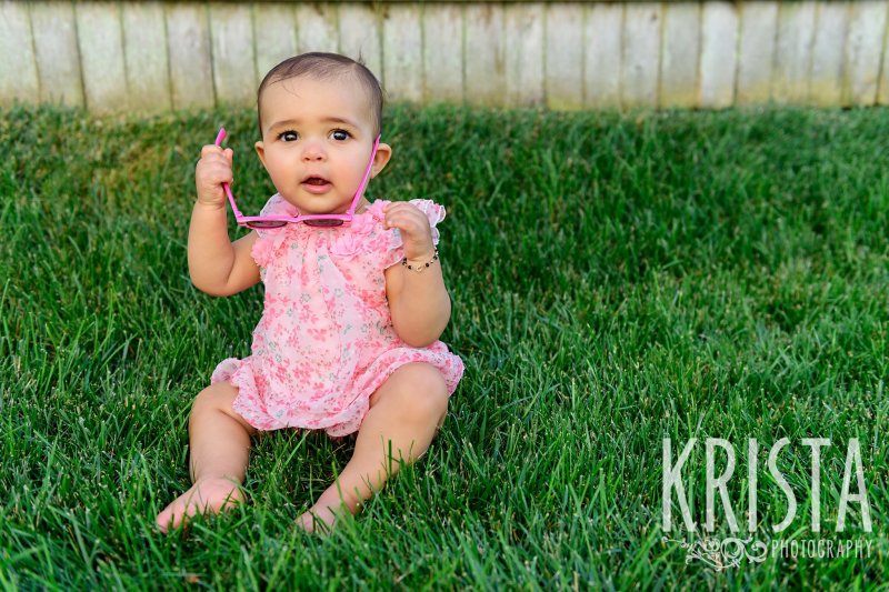 adorable baby girl in floral pink dress sitting on grass holding pink sunglasses during lifestyle portrait session at her family's home on Cape Cod