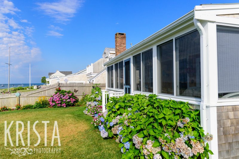 gorgeous Cape Cod home with blue sky and green grass and bushes around porch
