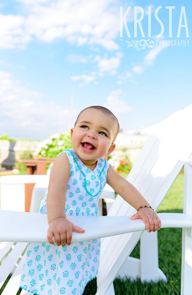 gorgeous happy baby girl standing on white adirondack chair in turquoise blue patterned dress