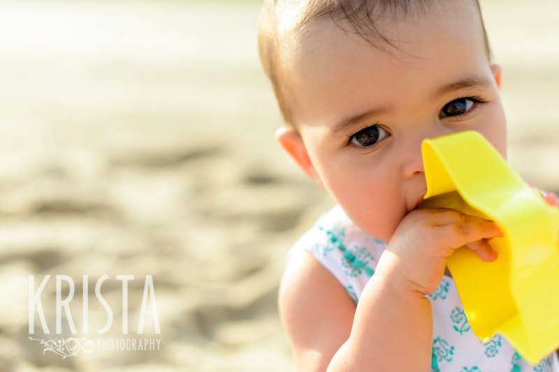 beautiful brown eyed baby girl chewing on sand toy on beach of Cape Cod during lifestyle portrait session 