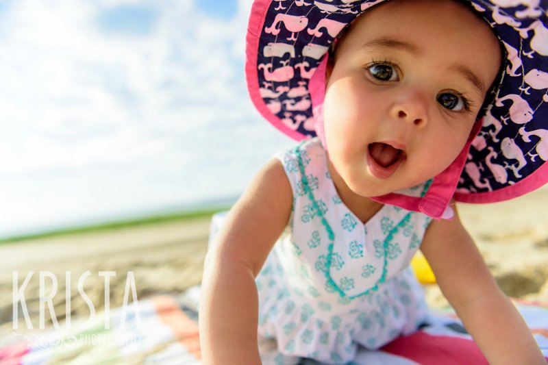 adorable baby girl in turquoise patterned dress with pink sun hat playing in sand of beach on Cape Cod during lifestyle portrait session