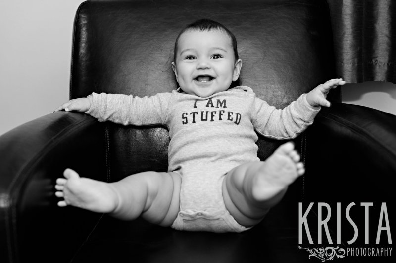 black and white image of six month old boy in leather chair wearing I am Stuffed onesie during lifestyle portrait session at family home