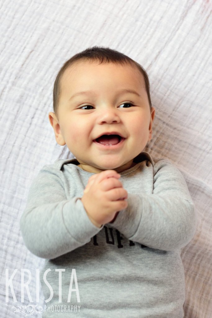 adorable six month old baby boy clasping hands together and smiling during lifestyle portrait session at family home