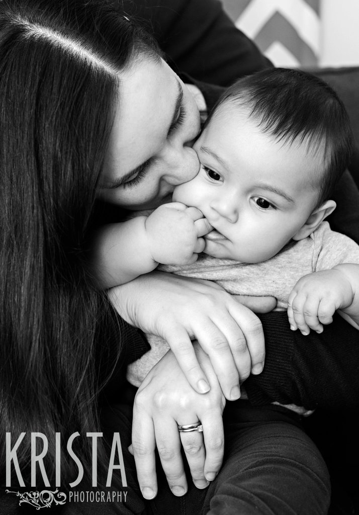 black and white image of mother kissing six month old baby boy on the cheek as he chews on a finger during lifestyle portrait session at home