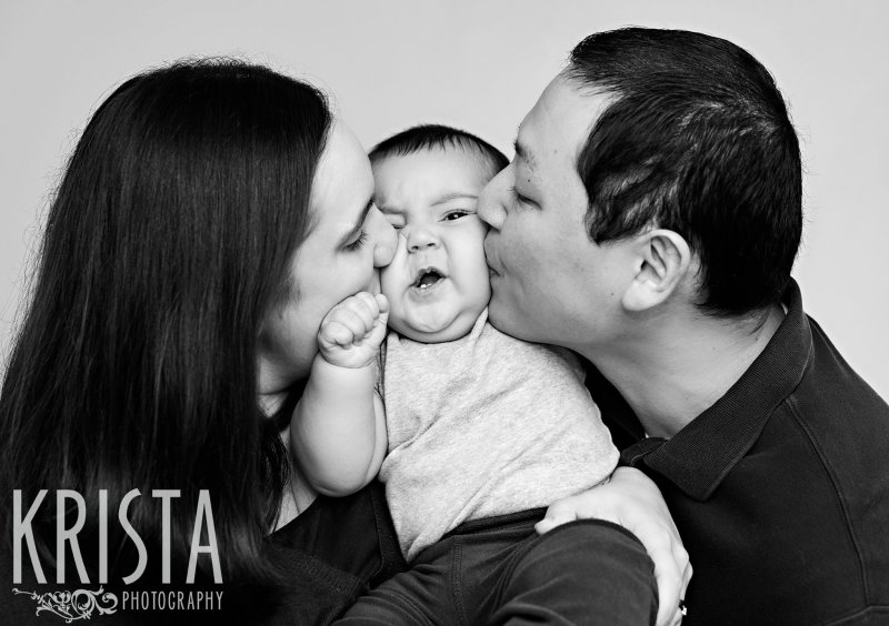 black and white image of six month old baby boy being kissed on both cheeks by adoring parents in big squishy family hug