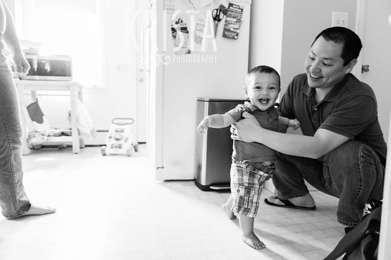 black and white image of one year old baby boy learning to walk with his dad in the kitchen