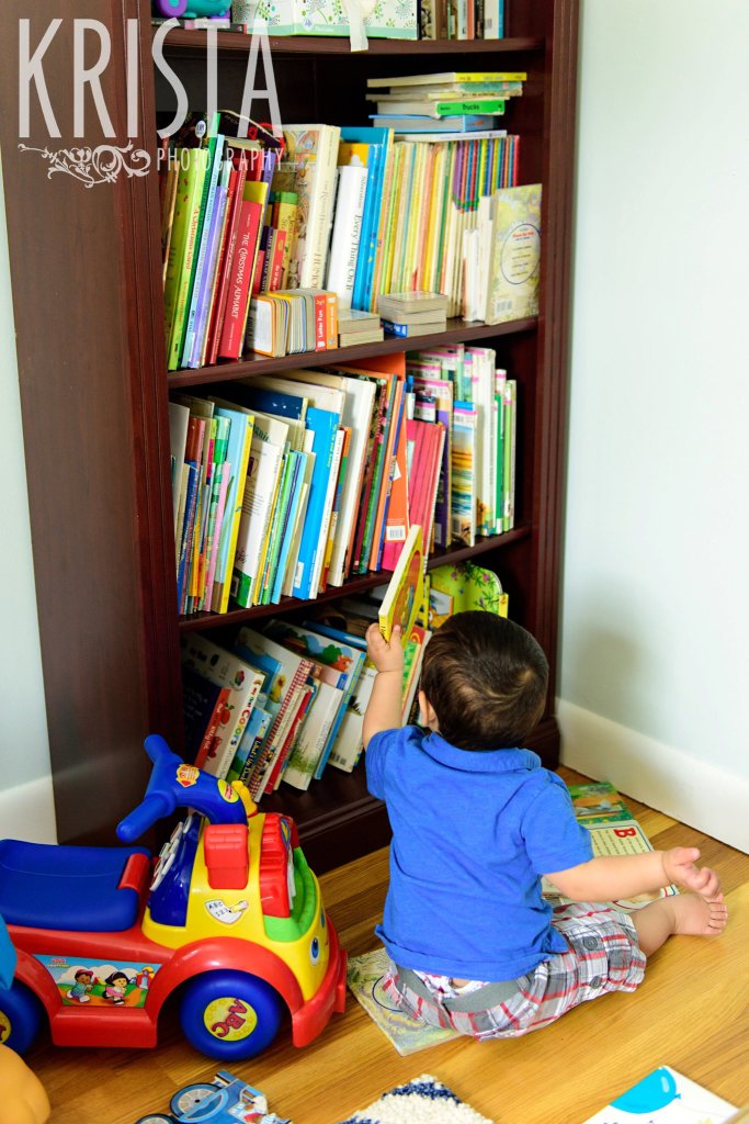 one year old baby boy in blue collared shirt taking book off of bookshelf during lifestyle portrait session at home