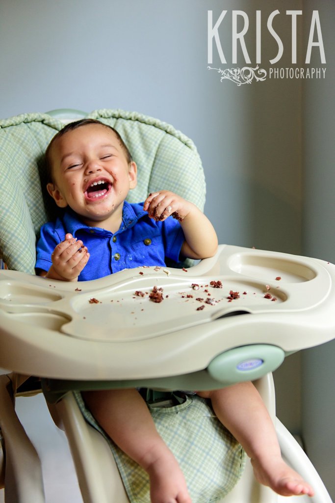 one year old baby boy laughing in his highchair while finishing first birthday cake during lifestyle portrait session at home