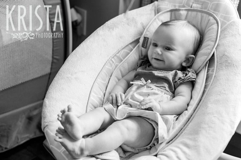 black and white image of three month old baby girl sitting in swing grinning with ankles crossed during lifestyle portrait session at home.