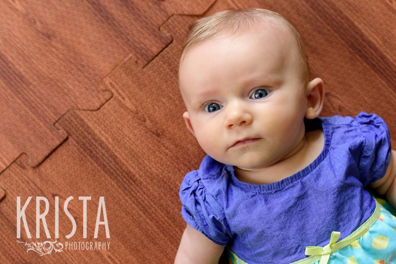 serious three month old baby girl laying on back on wood floor during lifestyle portrait session at home
