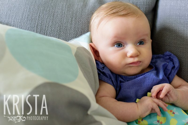 serious three month old baby girl propped up on blue polka dot pillow on gray couch during lifestyle portrait session at home
