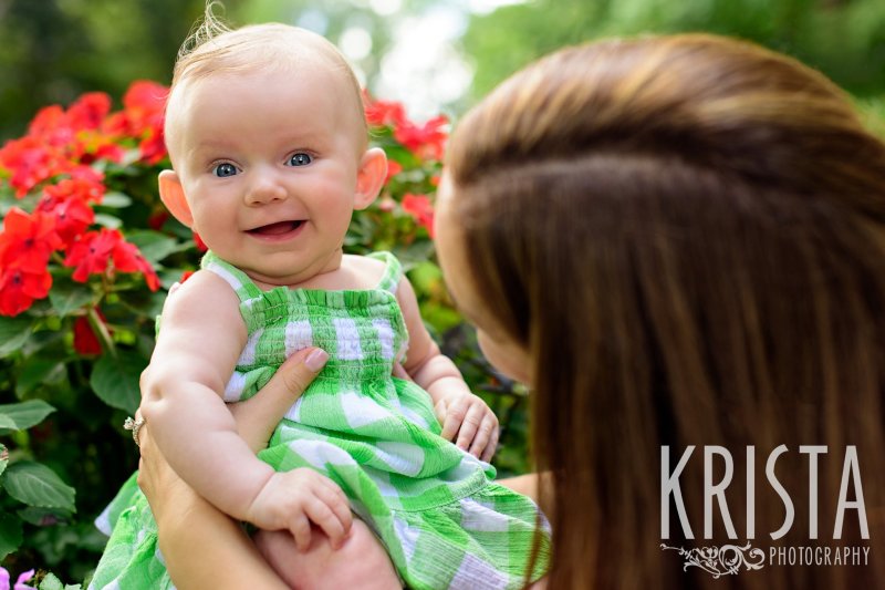 mother from behind holding three month old baby girl in green gingham sundress who is smiling during lifestyle portrait session at family home