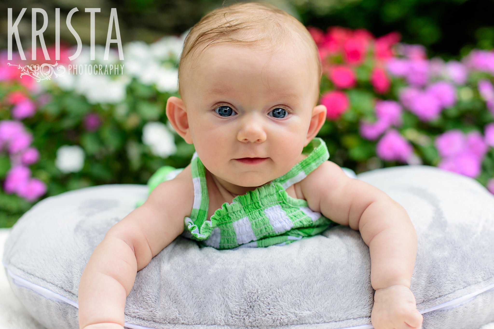 sweet little three month old baby girl in green gingham sundress laying on belly on pillow in front of pink and white flowers during lifestyle portrait session at home